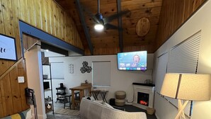 Un saló confortable amb un sofà gran, una llar de foc, una televisió i un ventilador de sostre.