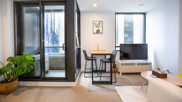 A modern living room with a sliding glass door leading to a balcony, a wooden dining table with chairs, a flat-screen TV, and a potted plant. - Cosy Apartment in the heart of Melbourne + Free Wi-Fi + Free Parking (Melbourne)