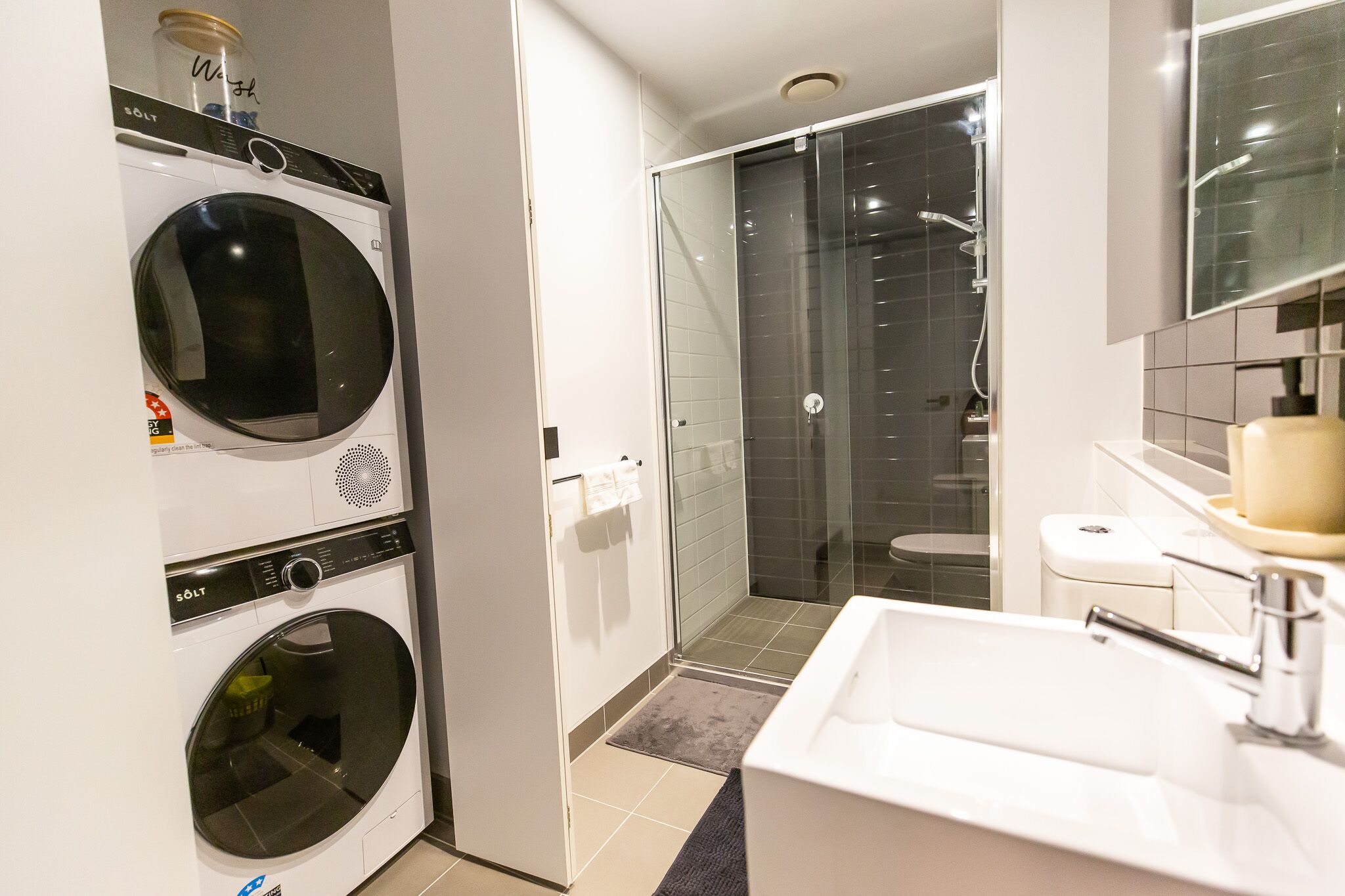 A modern bathroom with a built-in washer and dryer, a glass shower enclosure, and a white sink.