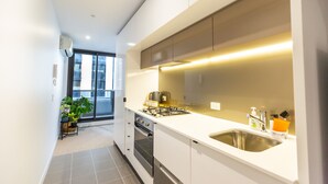 A modern kitchen with a gas stove, oven, and stainless steel sink. There is a window with blinds, a potted plant, and a small table with a basket. - Cosy Apartment in the heart of Melbourne + Free Wi-Fi + Free Parking (Melbourne)