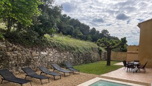Une piscine entourée d’un mur de pierre et de verdure, avec des chaises longues et une table à proximité.