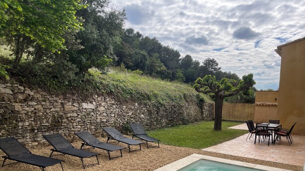 Una piscina con sillas de descanso y una mesa, rodeada por un muro de piedra y follaje.