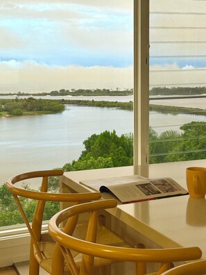 Una habitación con un amplio ventanal que da a un río, una silla de madera, una mesa con un libro y una taza.