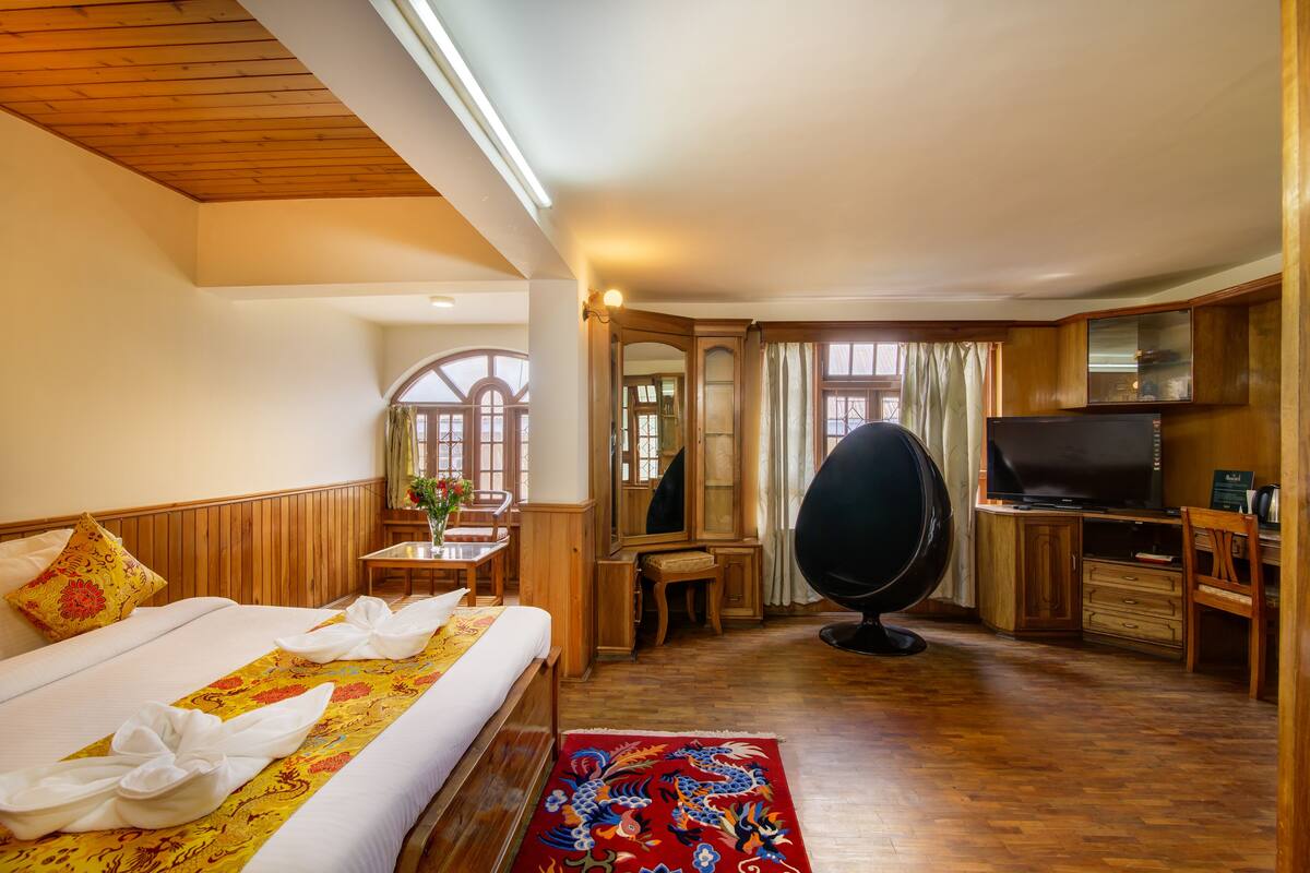 A hotel room with a wooden ceiling, a large bed with neatly made sheets, a desk with a chair, a television, and a large mirror.
