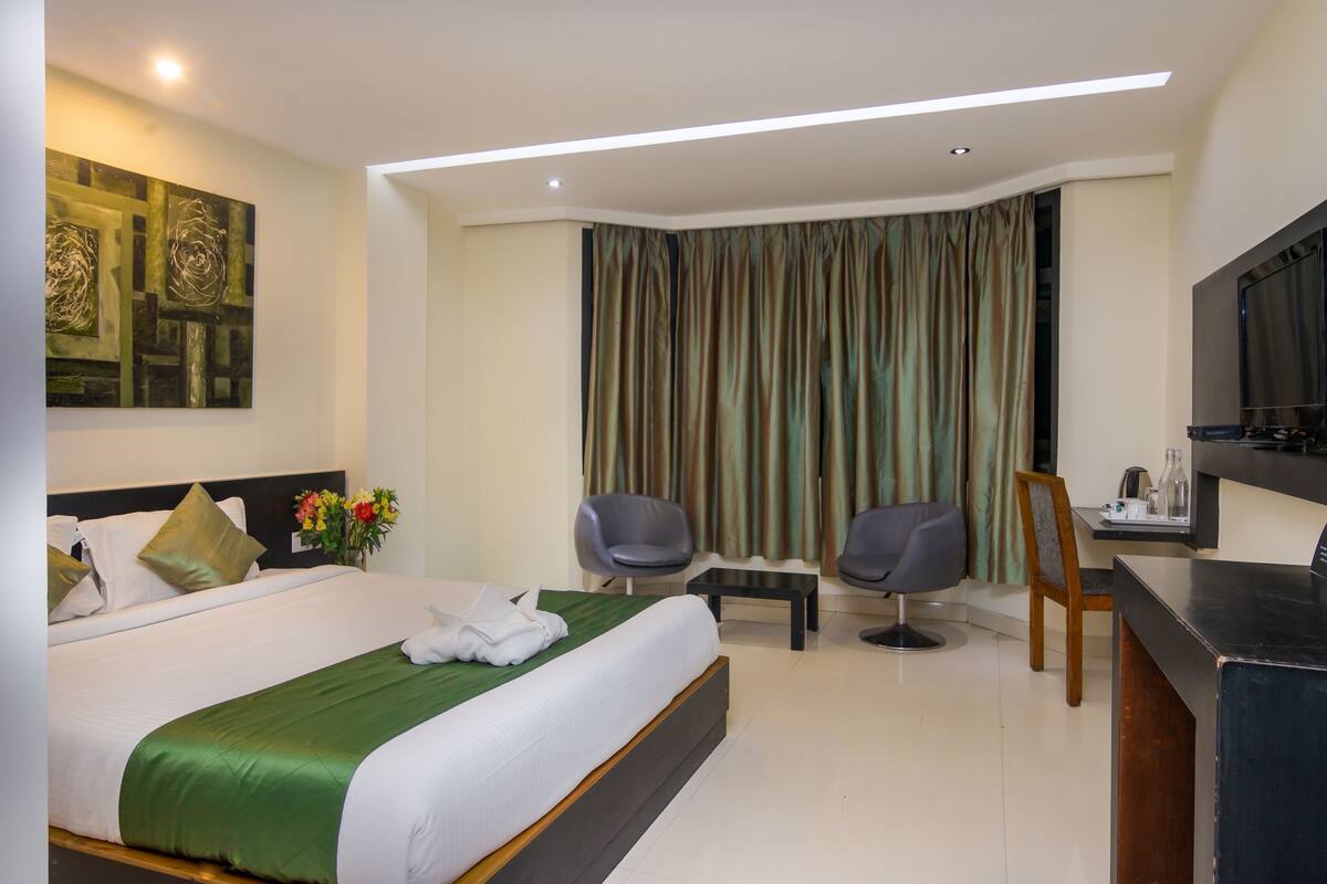 A hotel room with a bed, two chairs, a desk, a TV, and a painting on the wall.