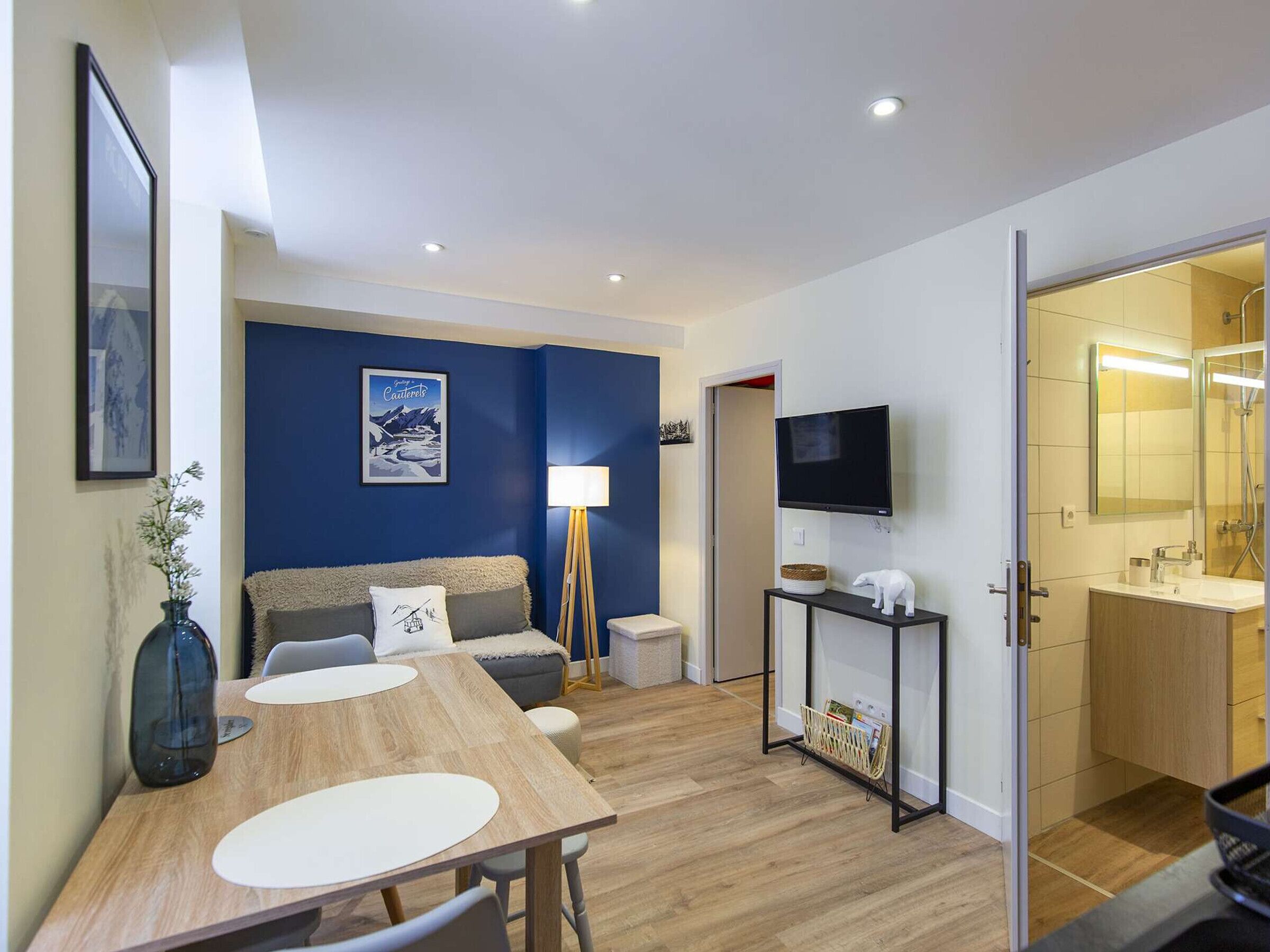 A modern living room with a blue accent wall, a wooden dining table, a sofa, a TV, and a small kitchen area with a mirror and sink.