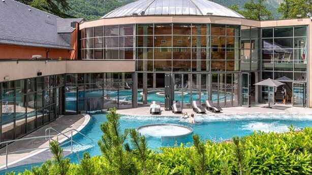 A hotel with a large glass-enclosed pool, outdoor seating, and a mountain backdrop. - Cozy 2-Room Apartment in Cauterets Center - Sleeps 2-4, Well-Equipped Kitchen, Near Ski Lifts (Cauterets)