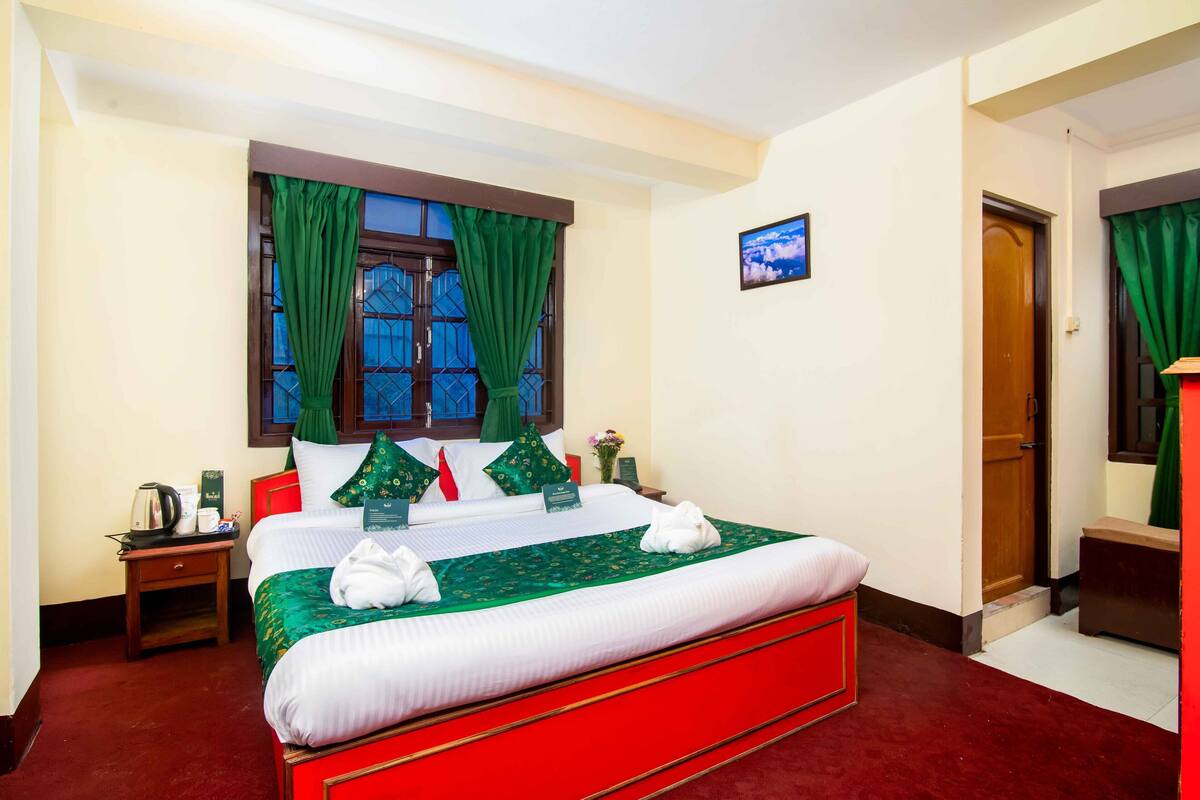 Una habitación de hotel con una cama grande, cortinas verdes, una mesita de noche de madera, una pequeña mesa con una tetera y un cuadro en la pared.