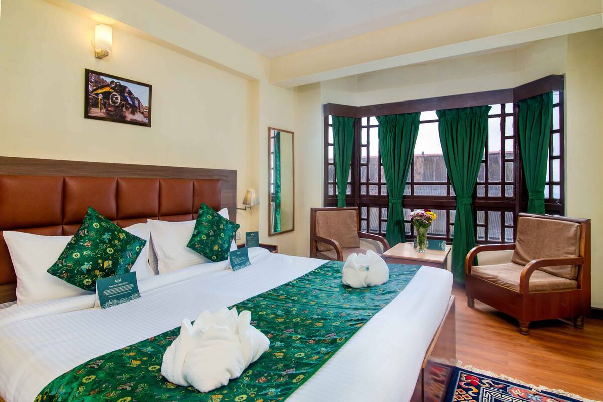 Habitación de hotel con una cama grande, dos sillas, un escritorio con un florero y una foto enmarcada en la pared, además de una ventana con cortinas verdes.