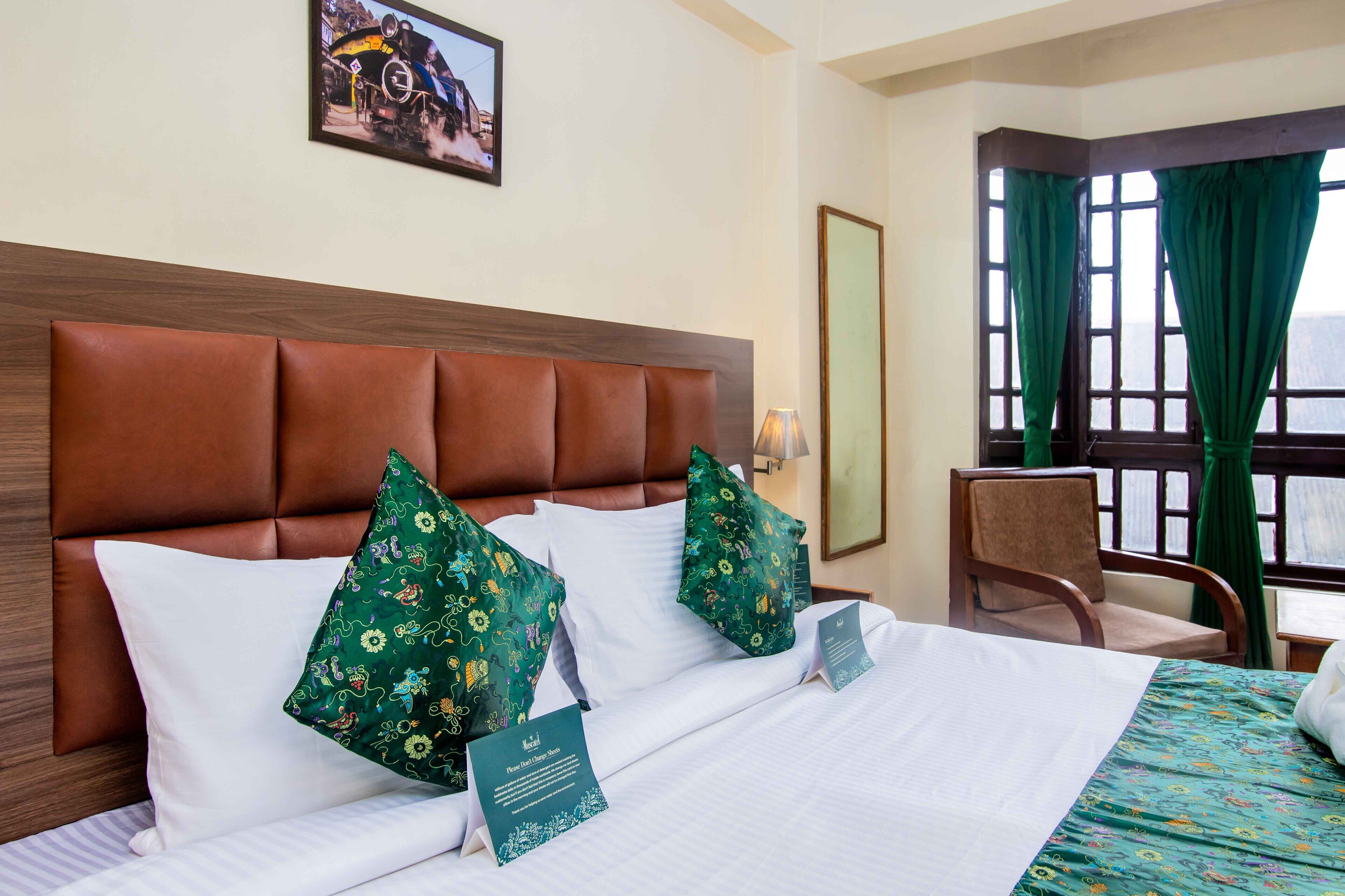 A hotel room with a brown leather headboard, floral-patterned pillows, a bed with white linens, a green floral bedspread, a framed picture on the wall, and a window with green curtains.