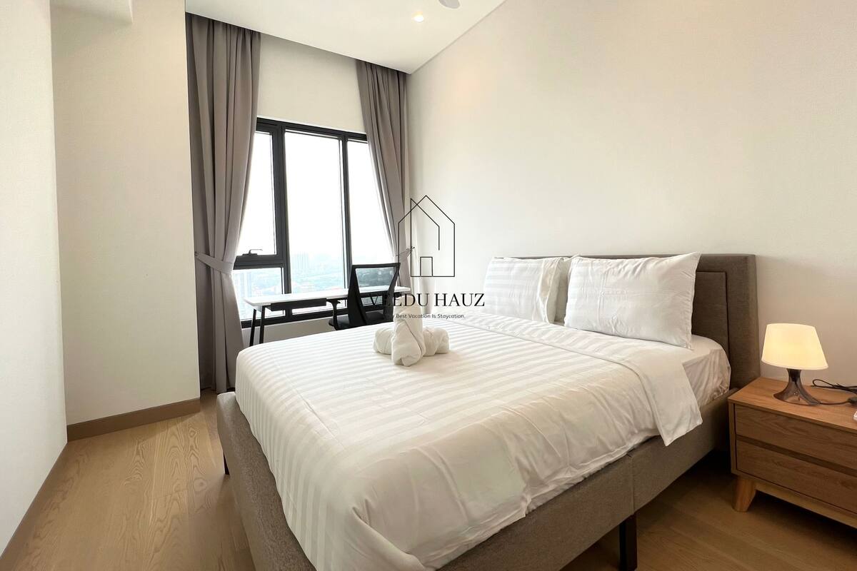 A modern hotel room with a large bed, a bedside table with a lamp, a ceiling fan, and a view of the city through large windows.