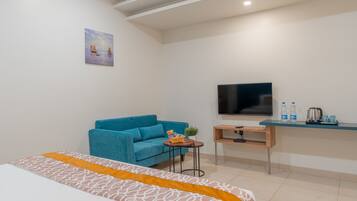 Quarto de hotel moderno com uma cama, um sofá azul, uma pequena mesa com uma planta, uma televisão e uma escrivaninha com uma chaleira e garrafas de água.