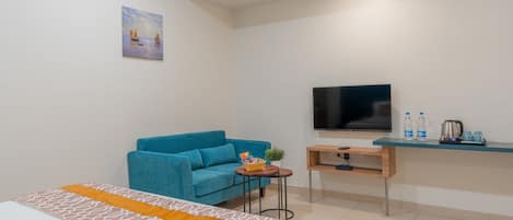 A modern hotel room with a bed, a blue sofa, a small table with a plant, a TV, and a desk with a kettle and water bottles.