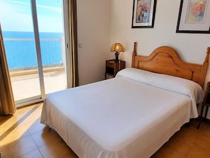 A bedroom with a bed, a nightstand, a lamp, and a view of the sea through a sliding door.