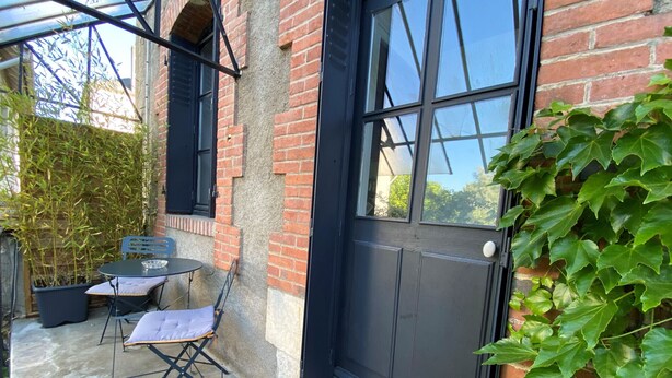 Un bâtiment en briques avec une porte et des fenêtres noires, un petit patio avec une table et des chaises, et une plante en pot.