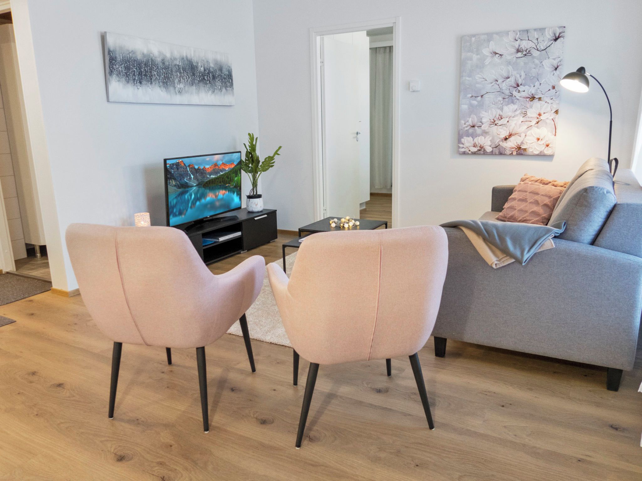 Un salon moderne avec un canapé gris, deux fauteuils roses, un téléviseur à écran plat et une plante.