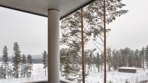 A view from a balcony with a metal railing, overlooking a snowy landscape with tall trees and a building in the distance.