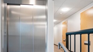A modern elevator with metallic finish, a wooden door, and a metal handrail in a hallway with tiled flooring.
