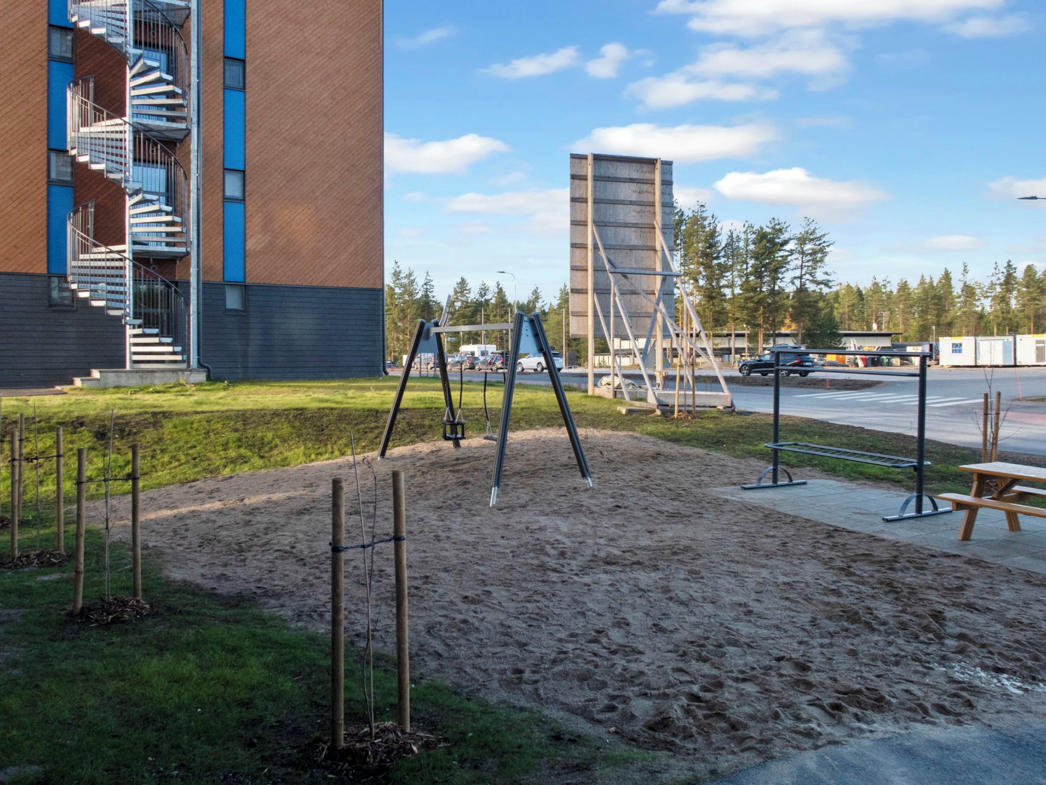 Leikkikenttä, jossa on keinut ja hiekkalaatikko. Kentän ympärillä on rakennus, jossa on portaikko, ja taustalla näkyy parkkialue.