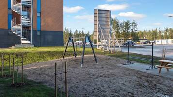 Leikkikenttä, jossa on keinut ja hiekkalaatikko. Kentän ympärillä on rakennus, jossa on portaikko, ja taustalla näkyy parkkialue.
