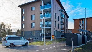 A modern multi-story building with balconies, a parking area, and a pedestrian crossing sign.