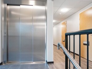 Un ascensor moderno con detalles metálicos, una puerta de madera a la derecha y un pasamanos metálico en primer plano.