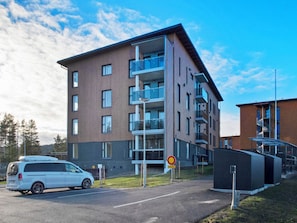 A modern multi-story residential building with balconies, a parking area, and a clear sky.
