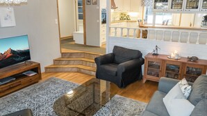 A living room with a TV, a sofa, a chair, a glass coffee table, and a staircase leading to a kitchen. - Torvokoski by Interhome (Utsjoki)
