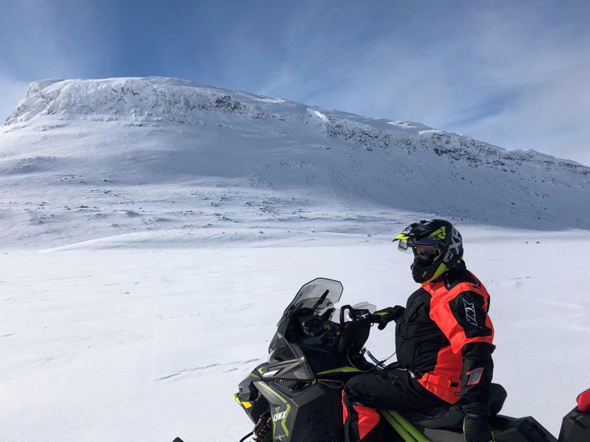 Ein Schneemobilfahrer in einer roten und schwarzen Ausrüstung, vor einer verschneiten Landschaft mit einem Berg im Hintergrund.
