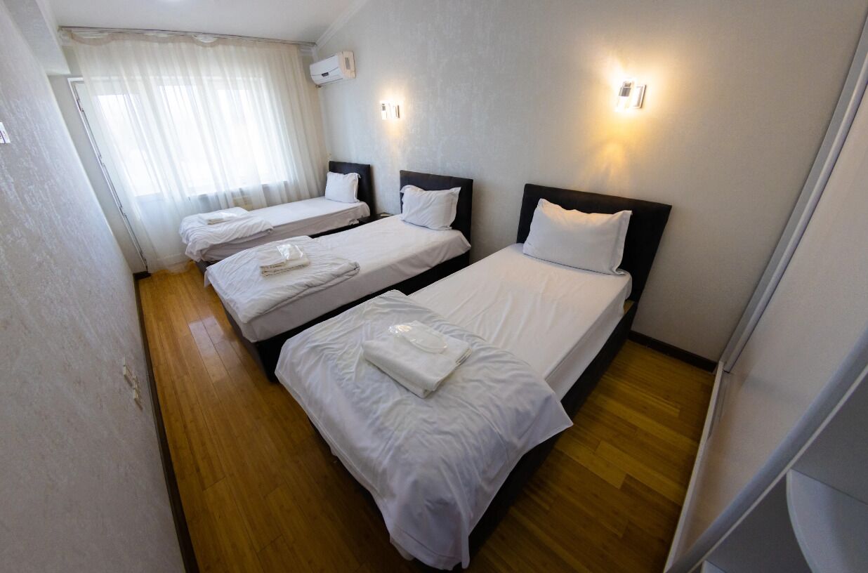 Une chambre d’hôtel avec trois lits simples, un plancher en bois, une fenêtre avec des rideaux légers et des luminaires fixés au mur.