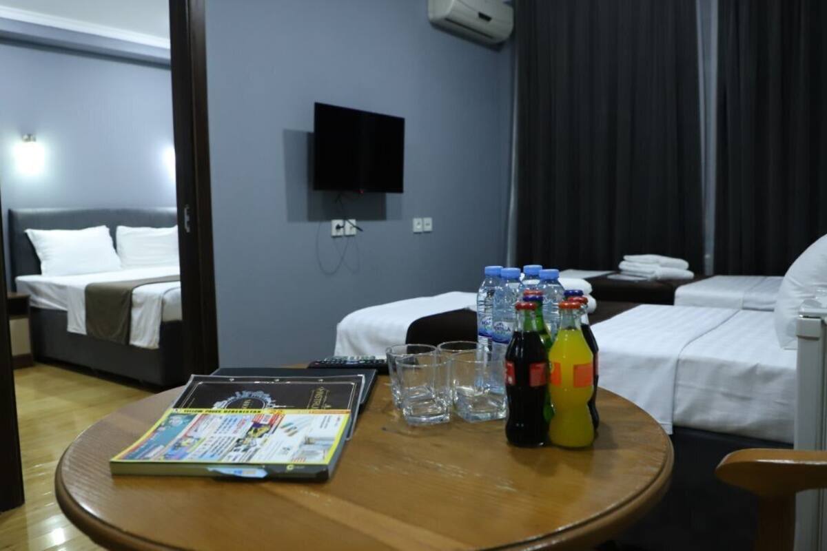 A hotel room with a bed, a TV mounted on the wall, a table with a magazine, glasses, and bottles of water and juice.