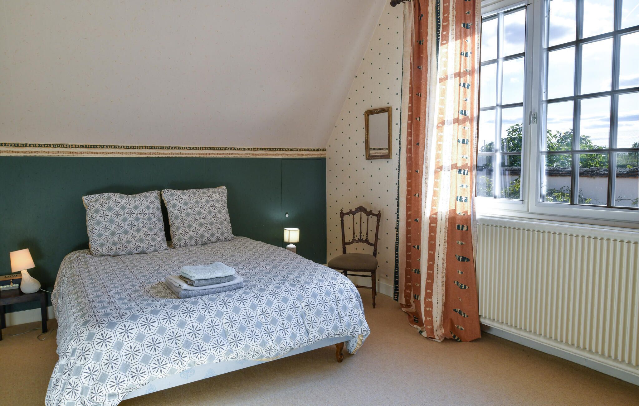 A bedroom with a bed, a chair, a nightstand, a lamp, a window with curtains, and a radiator.