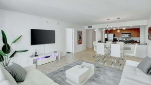 Un salon moderne doté d’un téléviseur à écran plat, de canapés blancs, d’une table basse et d’un coin repas avec des chaises, et une cuisine en arrière-plan.