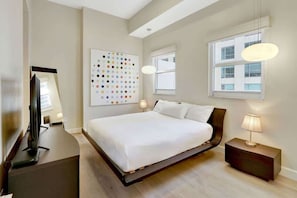 A modern hotel room with a large bed, a flat-screen TV, a wall art piece, and a window providing natural light.