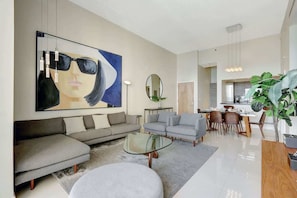 A modern living room with a large abstract painting, a sofa, armchairs, a glass coffee table, and a dining area with chairs and a plant.