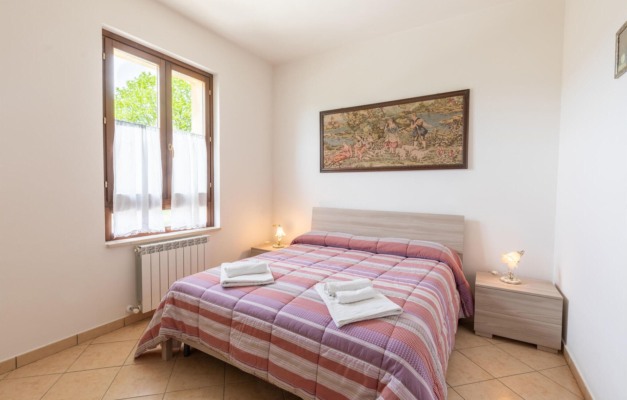 A bedroom with a bed, a nightstand, a window with curtains, a radiator, and a painting on the wall.