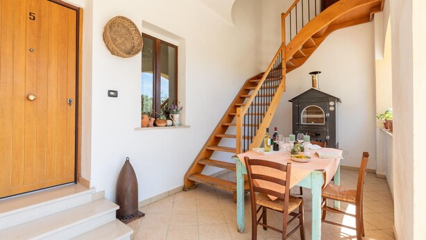 Un escalier en bois menant à un deuxième étage, une porte en bois rustique avec un numéro, une fenêtre ornée de plantes en pot et un coin cuisine avec un poêle.