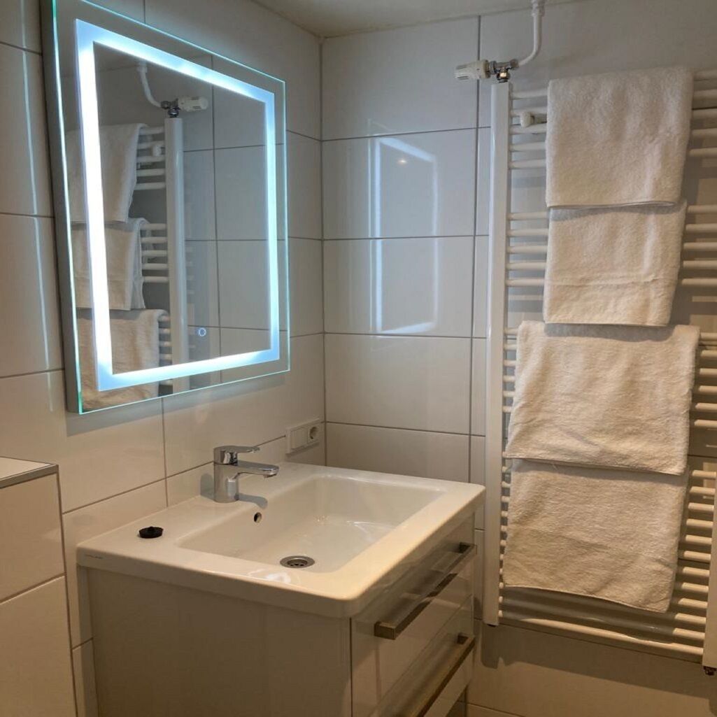 Un baño con un lavamanos blanco, un espejo con luz y un perchero con toallas.