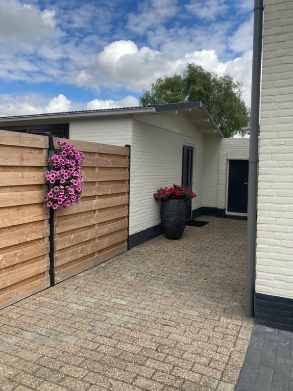 A covered outdoor area with a wooden fence, a white building with a black door, and a large planter with flowers.