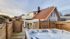 Une terrasse en bois mène à une maison dotée d’un toit en pente, d’un jacuzzi et offrant une vue sur le paysage environnant.