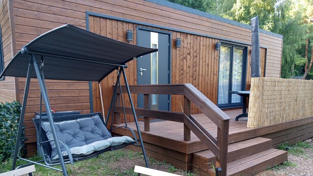 A wooden deck with a swing chair and a bench, leading to a modern building with large windows. - Tiny house in unspoiled nature on Lake Plau (Bad Stuer)