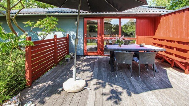 Eine Holzdielen-Terrasse mit Tisch und Stühlen, ein roter Zaun und ein überdachter Bereich mit Sonnenschirm.