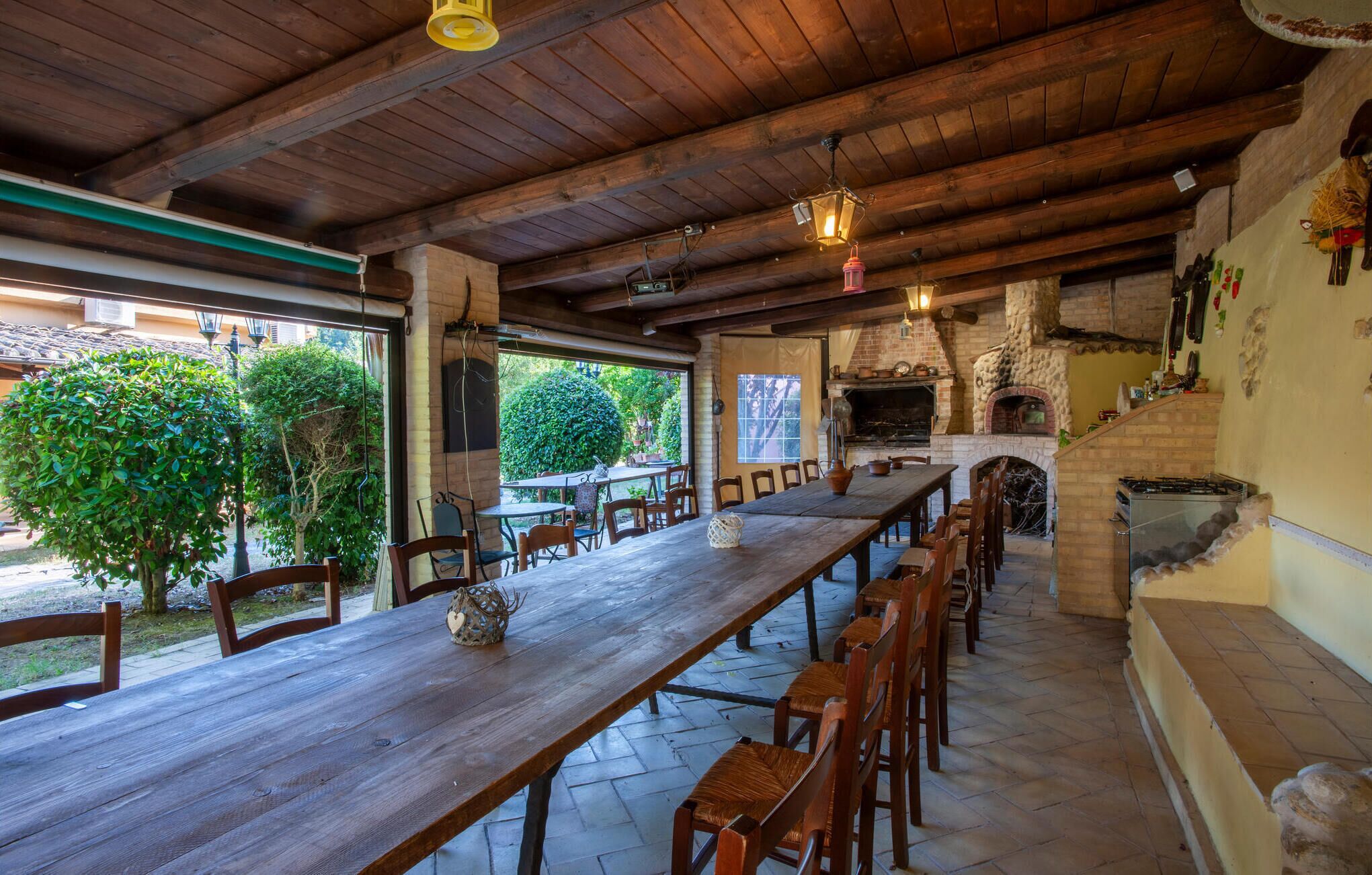A rustic outdoor dining area with a long wooden table and chairs, a brick oven, and a view of greenery.