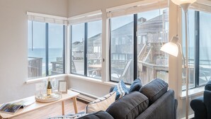 Une pièce chaleureuse avec une grande fenêtre donnant sur une terrasse en bois et l’océan. On y trouve un canapé, une table sur laquelle est posé un plateau avec divers objets, et un lampadaire.