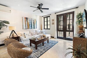Un salon spacieux doté d’une grande fenêtre, d’un ventilateur de plafond et d’un canapé agrémenté de coussins décoratifs.
