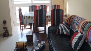 Een woonkamer met een zwarte leren bank, een houten salontafel, een eettafel met stoelen en een kroonluchter.