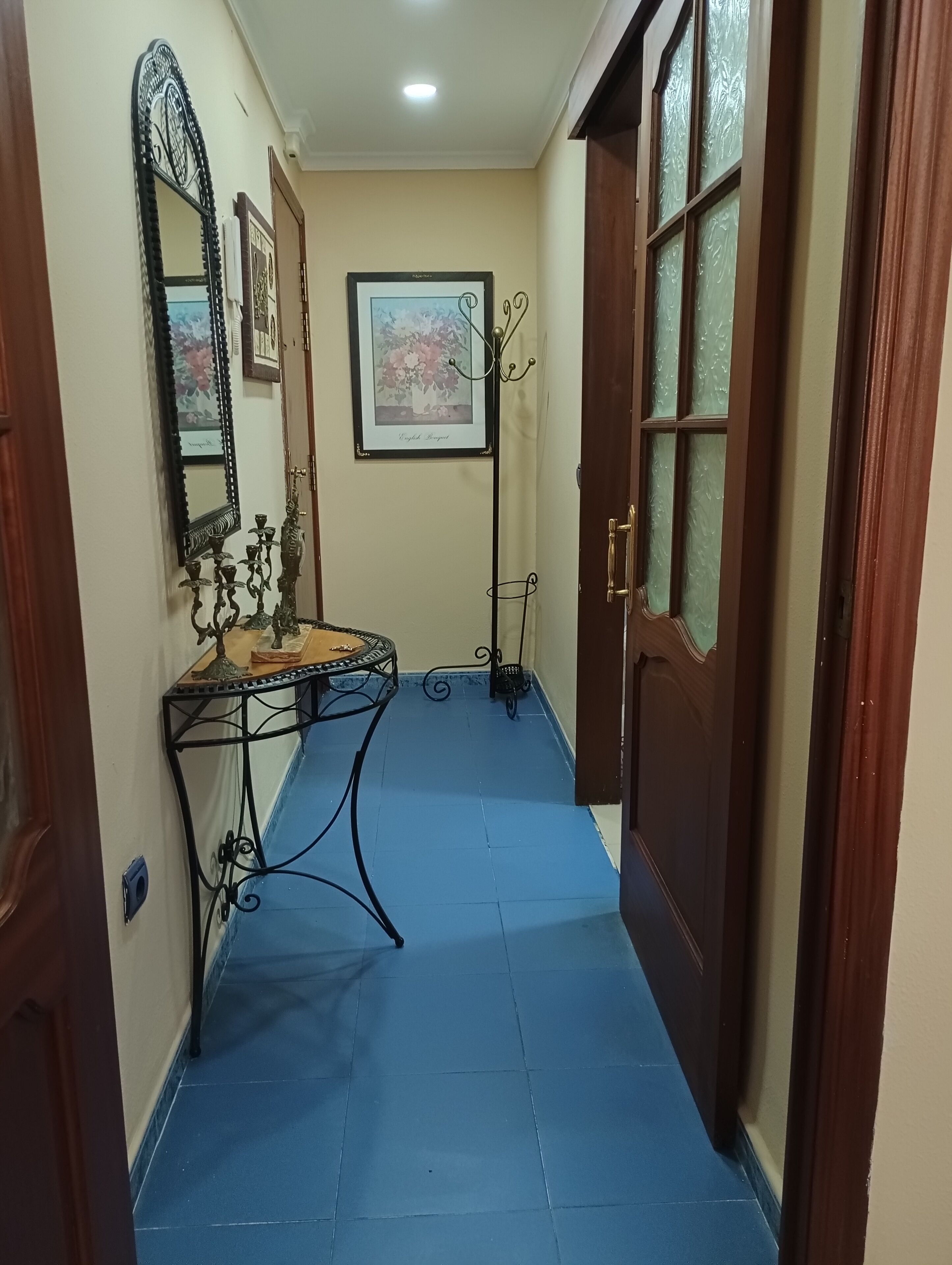 Een smalle gang met een vloer van blauwe tegels, een houten deur met ruitpanelen, een wanddecoratie met bloemen en een klein tafeltje met spiegel en kandelaars.