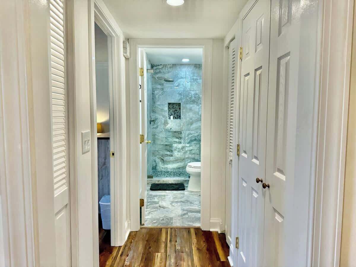 A hallway leading to a bathroom with a marble shower wall, a toilet, and a wooden floor.