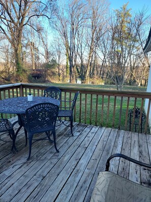 A wooden deck with a table and chairs, surrounded by a railing, overlooking a grassy yard with trees.