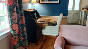 Una habitación con piso de madera, un sofá rosa, un escritorio con una computadora portátil, una lámpara y un cuadro en la pared.
